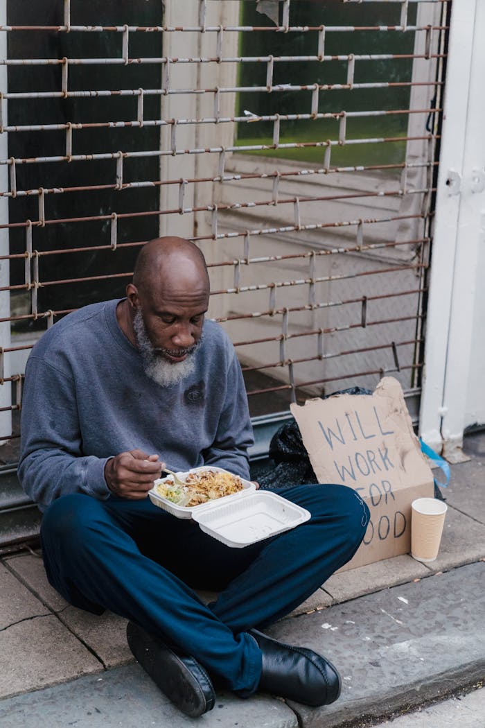 A homeless man sitting on the sidewalk eating a meal with a cardboard sign beside him, highlighting social issues.